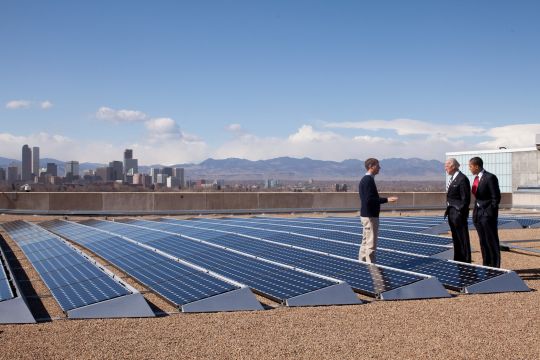 Barack Obama und Joe Biden unterhalten sich auf einem Solardach mit Blake Jones, dem Chef von Namaste Solar Electric, im Hintergrund Hochhäuser von Denver.