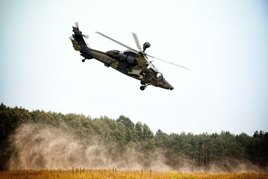 Armeehubschrauber fliegt schräg über einem Feld, viel Staub wird aufgewirbelt.