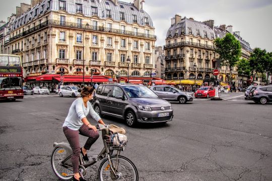 Stadtplatz mit Autos und einer E-Bike-Fahrerin, im Hintergrund französische Altbauten.