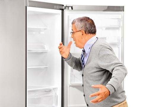 Älterer Mann in Hemd und Weste schaut in den Kühlschrank und erschrickt, weil er leer ist.