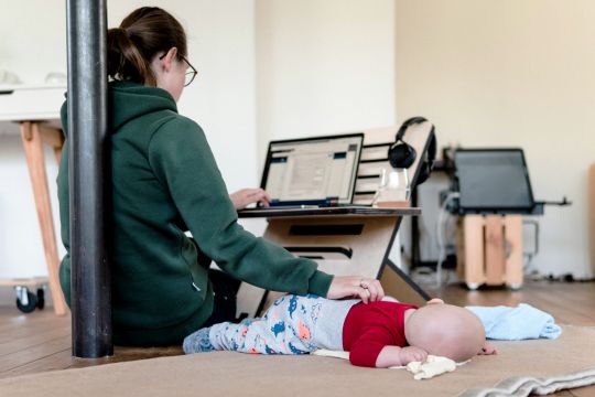 Junge Frau arbeitet in minimalistisch eingerichtetem Zimmer auf dem Boden am Laptop, neben ihr liegt ein Baby auf einer Matte.