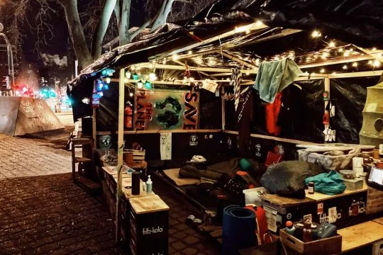 Selbstgebaute Klimacamp-Hütte aus Holzstangen, Kisten und Plastikplanen, mit Schlafsäcken, Proviant und Plakaten, bei Nacht aufgenommen.
