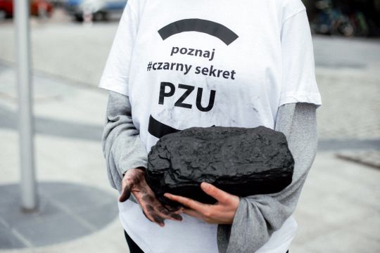Weiß gekleidete Person hält ein großes Stück Kohle in den geschwärzten Händen, auf dem T-Shirt ist auf Polnisch die Aufschrift: Das dunkle Geheimnis lüften – PZU.