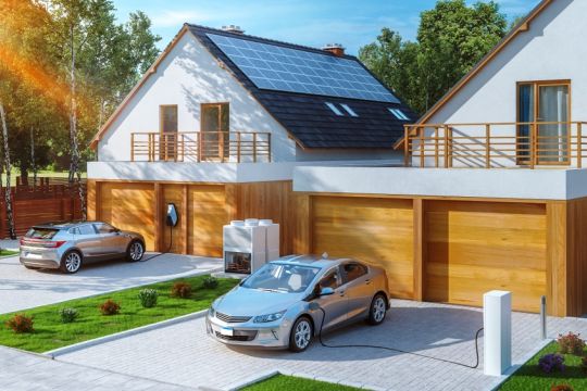 Zwei neue Einfamilienhäuser mit Solardach und je zwei Garagen, davor steht jeweils ein Elektroauto, das gerade aufgeladen wird.