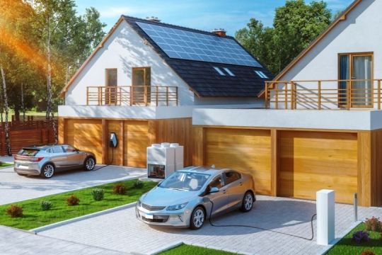 Zwei neue Einfamilienhäuser mit Solardach und je zwei Garagen, davor steht jeweils ein Elektroauto, das gerade aufgeladen wird.