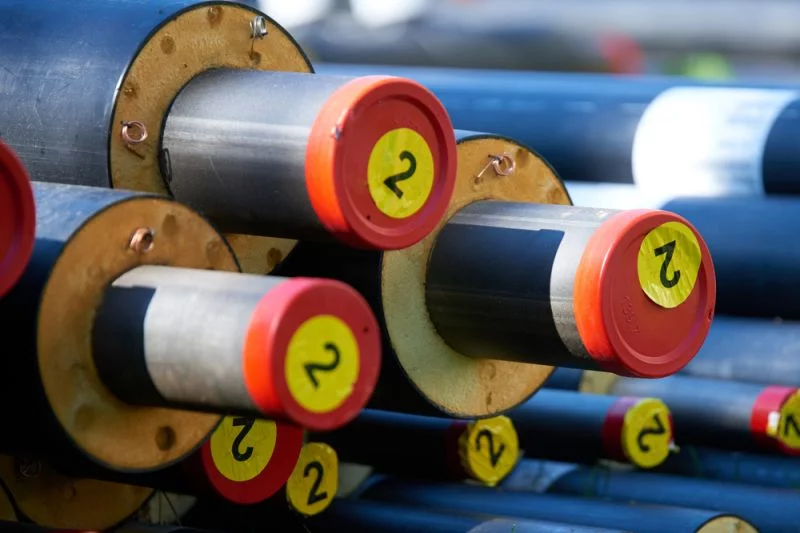 Neue Fernwärme-Rohrleitungen mit den typischen roten Schutzabdeckungen liegen auf einem Stapel, alle sind mit einem runden gelben Aufkleber gekennzeichnet, auf dem die Zahl 2 steht.