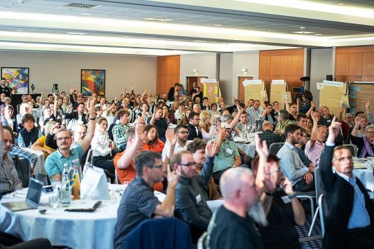 Etwa hundert Menschen sitzen in einem Saal verteilt an Tischen, aber alle in eine Richtung gewandt, ungefähr die Hälfte hebt den Arm zur Abstimmung.