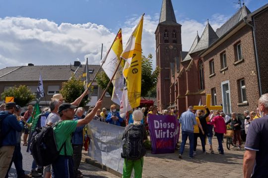 Menschen vor der Kirche in Keyenberg mit bunten Fahnen, gelben Kreuzen und Transparenten wie: Diese Wirtschaft tötet.