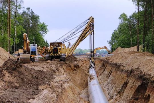 Verlegung einer Pipeline mit Baggern und Kränen in einen ausgebuddelten Graben in einer Schneise im Wald.