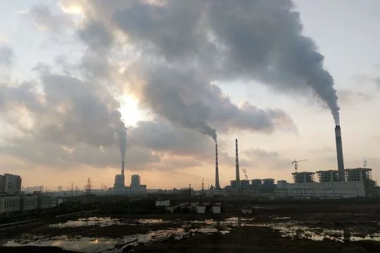 Großes Kohlekraftwerk mit mehreren stark rauchenden Schornsteinen, davor eine teils unter Wasser stehende Brachfläche.