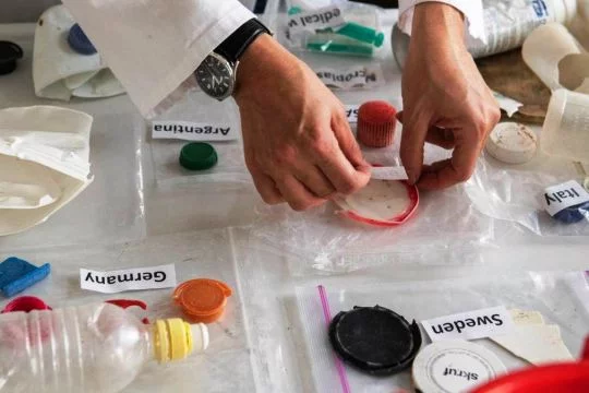Auf durchsichtige Tüten mit Plastikmüll, der schon lange im Meer war, werden Schildchen mit englischen Ländernamen geklebt: Schweden, Deutschland, Argentinien, Italien.