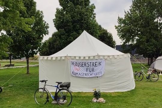 Auf einer Wiese ein rundes weißes Zelt mit der Aufschrift: Hungerstreik für Klimagerechtigkeit. Davor ein Fahrrad.