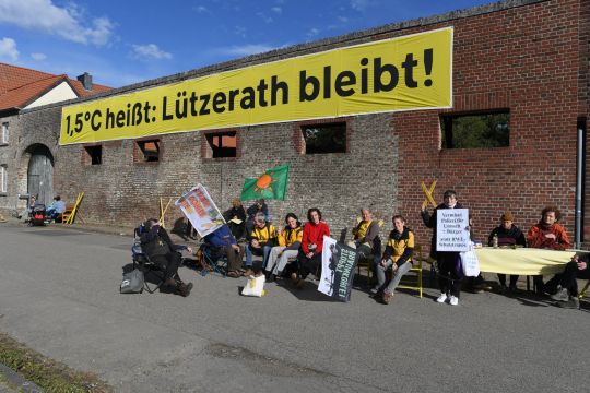 Eine Gruppe von Protestierenden sitzt vor einer Mauer mit einem Banner: 1,5 Grad heißt: Lüzerath bleibt!