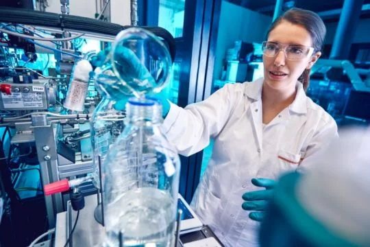 Labormitarbeiterin mit Schutzbrille gießt farblose Flüssigkeit in durchsichtige Flasche..