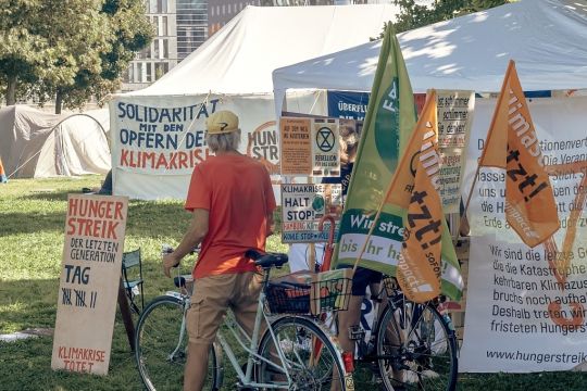 Ein Mann mit Fahrrad steht vor dem weißen Leinwandzelt der Hungerstreikenden, etliche Transparente und Fahnen zur Unterstützung sind zu sehen.