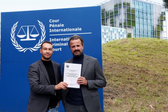 Zwei Männer stehen vor dem Gebäude des Internationalen Strafgerichtshofs in Den Haag und halten eine Klageschrift in der Hand.