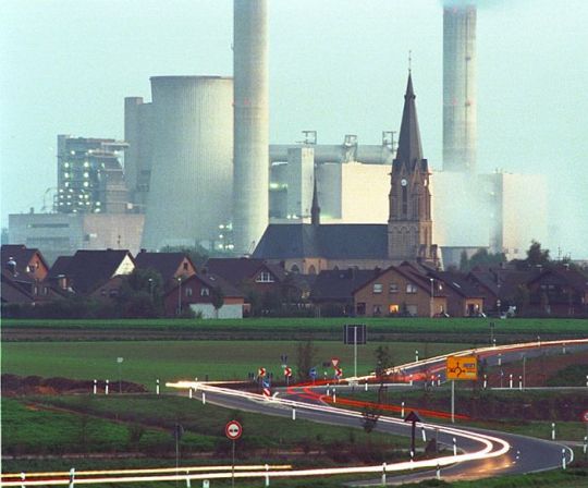 Dorf mit hinführender Straße, dahinter ein riesiges Kohlekraftwerk.