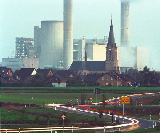 Dorf mit hinführender Straße, dahinter ein riesiges Kohlekraftwerk.