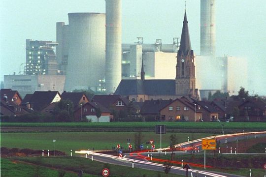 Dorf mit hinführender Straße, dahinter ein riesiges Kohlekraftwerk.