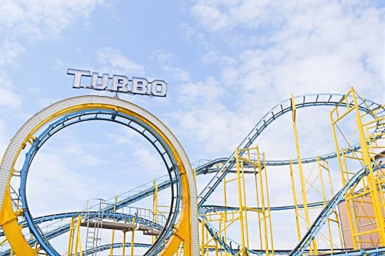 Vor leicht bewölktem Sommerhimmel eine blau-gelbe Achterbahn mit dem Schriftzug: Turbo.