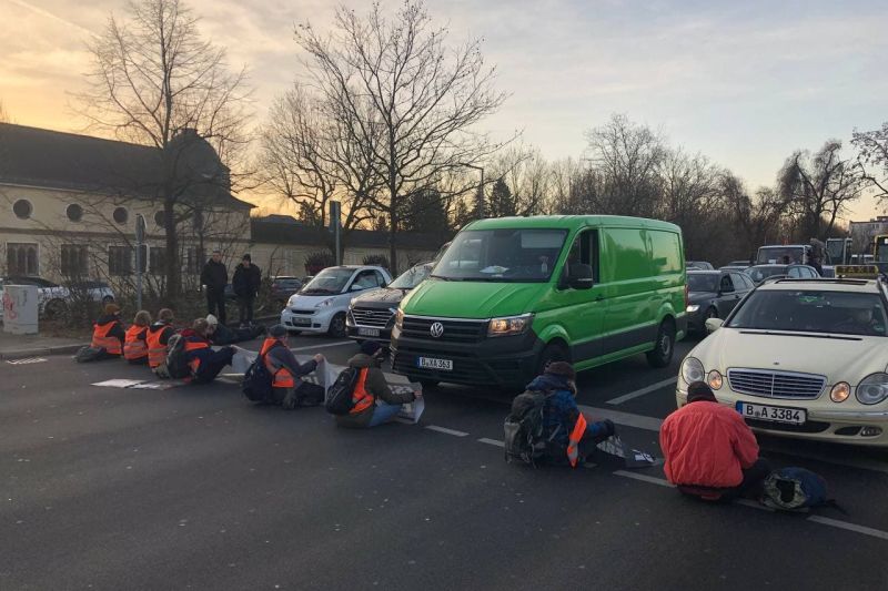 Mehrere Aktivist:innen in Warnwesten blockieren sitzend vier Spuren einer Straße