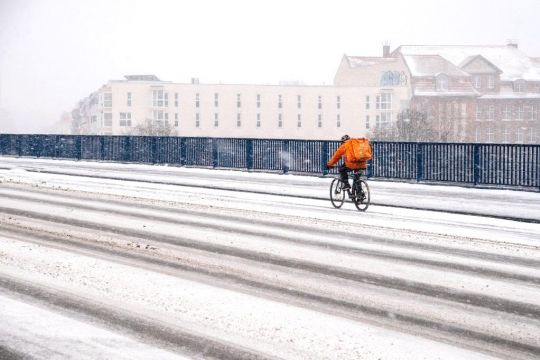 Ein einsamer Essenslieferant in Orange radelt auf einer langen verschneiten Brücke durch die Stadt.