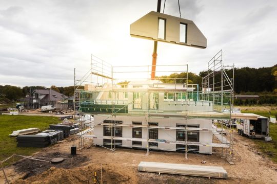 Baustelle eines Einfamilienhauses auf der grünen Wiese, der Kran schwenkt gerade ein Wandelement für die obere Etage ein.