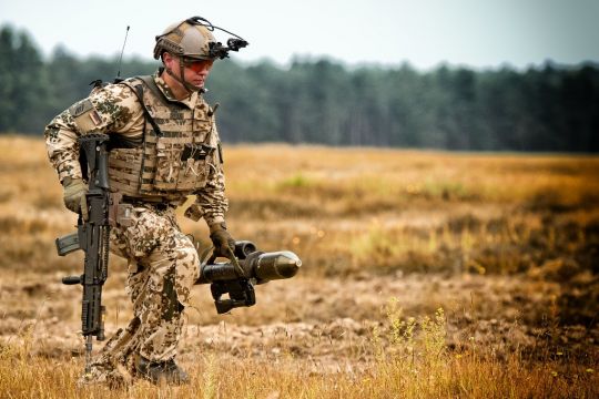 Ein Mann in beige-braun-grün geflecktem Tarnanzug läuft schnell über ein Feld, in der Hand hält er eine Pazerfaust und ein Gewehr, auf dem Kopf hat er einen Spezialhelm mit Kamera.