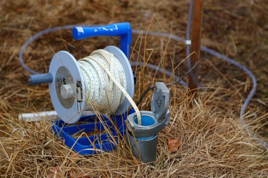 Grundwasserstandsmessung: Ein Messband wird in ein senkrecht aus dem im Boden ragendes Rohr eingelassen.