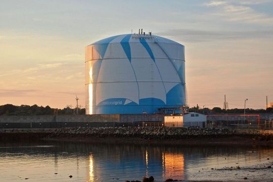 Zylinderförmiger, hellblau-weißer Gastank von der Größe eines mehrstöckigen Hauses in der Dämmerung, unten die Aufschrift National Grid, davor ein Gewässer, in dem sich der Tank spiegelt.