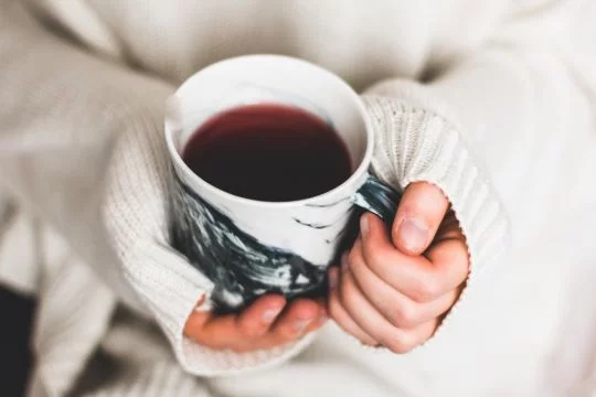 Aus einem weißen, warmen Pullover schauen nur die Hände hervor, die eine Tasse Tee halten.