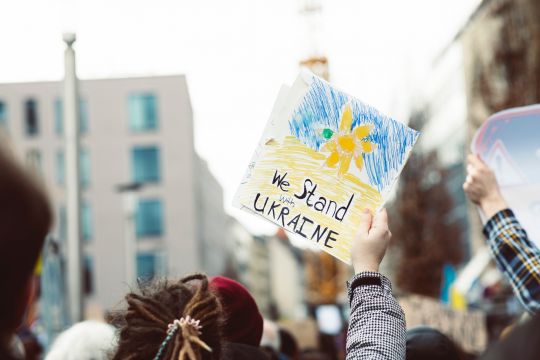 Selbstgemaltes blau-gelbes Schild auf einer Demo: We stand with Ukraine - Wir stehen der Ukraine bei.