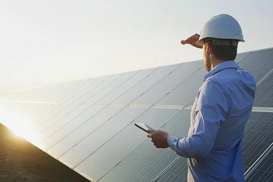 Neben einer langen, schräg aufgeständerten Solarstromanlage steht ein Mann im Hemd mit Helm und Tablet und schaut unter dem Schutz der Hand gegen die Sonne.