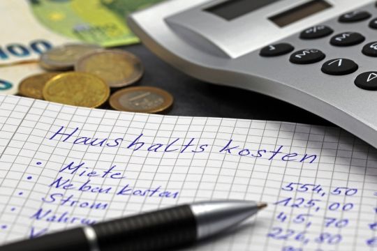 Liste der Ausgaben eines privaten Haushalts in deutscher Sprache auf Notizblock mit Stift, Münzen und Taschenrechner.