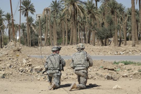 Zwei Soldaten in Tarnuniform knien neben einem Trümmerhaufen vor einem Palmenwäldchen.