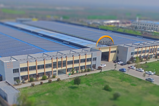 Große Fertigungshalle, über dem Eingang steht AE Solar mit dem Firmenlogo, davor einige Menschen und Autos.