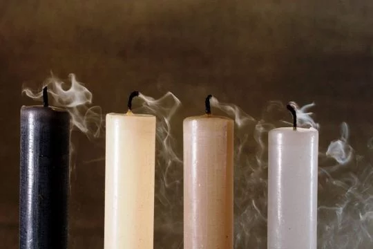 Vier Kerzen in Farben zwischen beige und schwarz vor dunkelbraunem Hintergrund, die gerade ausgeblasen wurden.