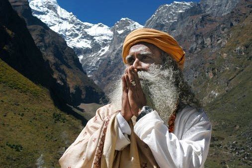 Jagadish Vasudev, genannt Sadhguru, steht in einem Hochgebirgstal, faltet die Hände und schaut etwas schräg nach oben. Der bärtige ältere Mann trägt einen weißen Mantel, ein helles, besticktes Übergewand und einen orangefarbenen Turban.