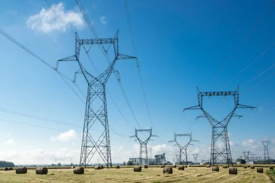 Mehrere Hochspannungsmaste auf einem Feld bei heiterem Himmel, am Horizont ein Kraftwerk.