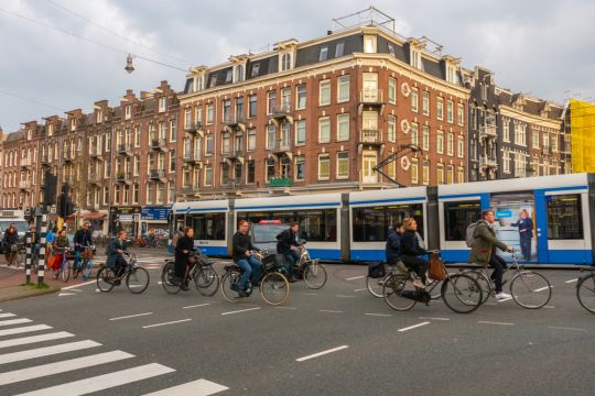Auf Hollandrädern fährt ein Dutzend Menschen über eine Kreuzung in einem Amsterdamer Altstadtviertel, eine blau-weiße Straßenbahn überholt sie.