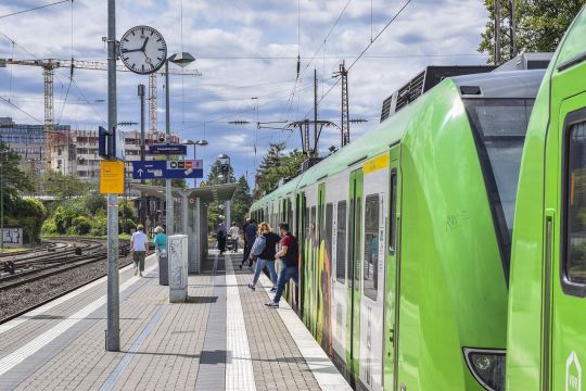 Grüner S-Bahn-Zug im Ruhrgebiet, einige Leute steigen um die Mittagszeit an einem nicht überdachten Bahnsteig aus, das Wetter ist sommerlich.