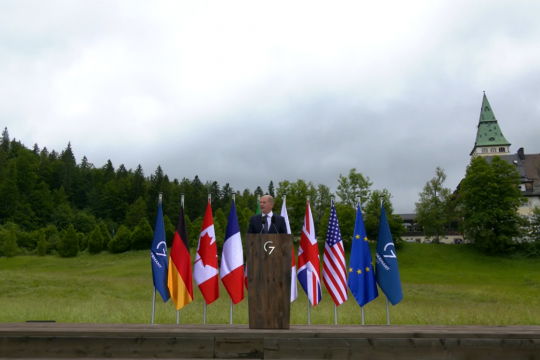 Bundeskanzler Olaf Scholz steht allein an einem Redepult, neben ihm die Fahnen der G7-Staaten, hinter ihm eine Wiese, rechts im Hintergrund Schloss Elmau.