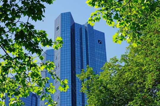 Doppelhochhaus der Deutschen Bank in Frankfurt am Main, am Bildrand überall das frische Grün sonnenbeschienener Bäume.