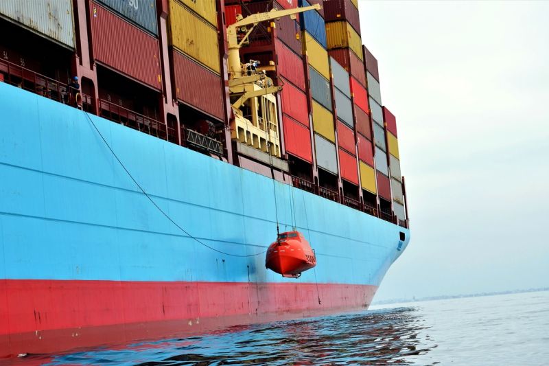 Containerschiff, an dem außenbords ein rotes Rettungsboot an einem Kran-Arm hängt.
