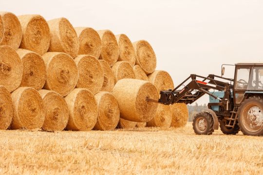 Auf einem Feld stapelt ein Traktor Strohballen mit einer Gabel.