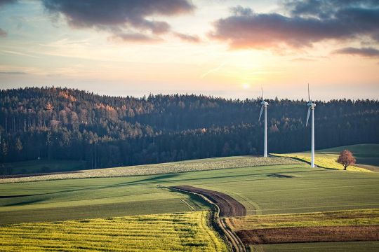 Zwei Windräder in hügeliger Landschaft mit Feldern und Wäldern.