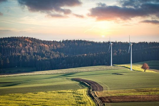 Zwei Windräder in hügeliger Landschaft mit Feldern und Wäldern. 