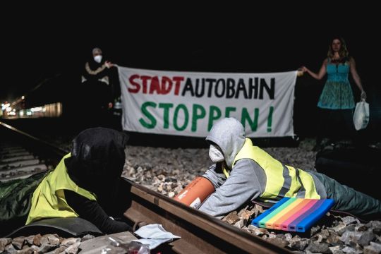 Zwei Klimaaktivistinnen haben sich an ein Bahngleis gekettet, im Hintergrund halten zwei weitere ein Transparent: Stadtautobahn stoppen!