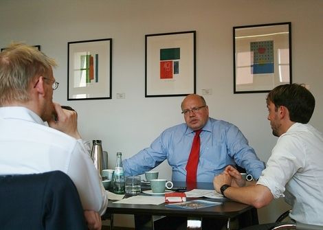 Peter Altmaier sitzt im hellblauen Hemd mit roter Krawatte am Tisch und stützt breit die Arme auf. Die Interviewer sind von schräg hinten zu sehen, beide im weißen Hemd.