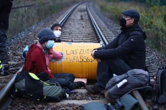 Gleisblockade mit einer gelben Tonne und zwei Menschen, die sich daran festgekettet haben.